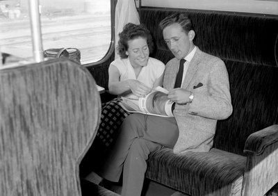 152860 Afbeelding van twee reizigers in een trein, met het tijdschrift Tussen de Rails van de N.S.